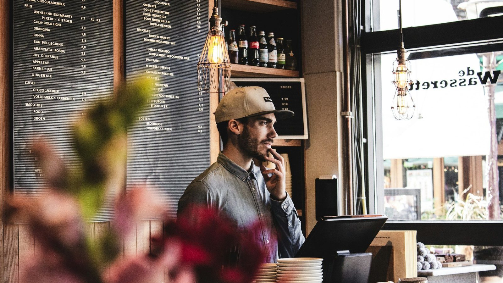 Cafe owner standing at the counter in front of the menu boards, thinking, with the register screen beside him